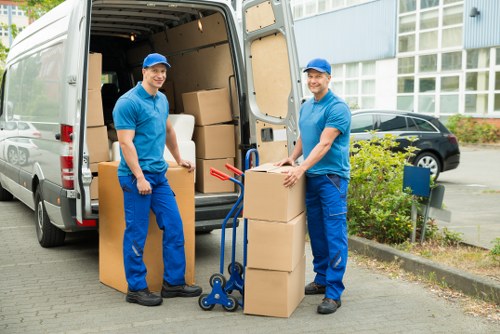 Two male removals workers dressed in matching blue uniforms and caps are standing outside a white removals van, which is parked on a paved driveway near a residential building with large windows. They are positioned on either side of a stack of cardboard moving boxes; one man is holding a box while the other is using a blue hand truck to transport another box. The boxes are of various sizes, some wrapped with packing tape, and are arranged in front of the open van door. Inside the van, additional boxes are visible, stacked on the floor. In the background, there are parked cars, green shrubbery, and a modern building with a light blue exterior. The scene appears well-lit under natural daylight, illustrating a professional house removal process in Greenwich, with a focus on careful handling and transportation of household items during a residential move.